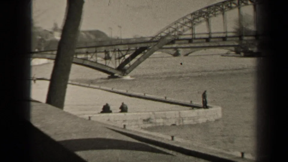 1947 Paris footage frame showing high water under a steel bridge, with the view partly blocked by tree trunks and small figures on the quay.