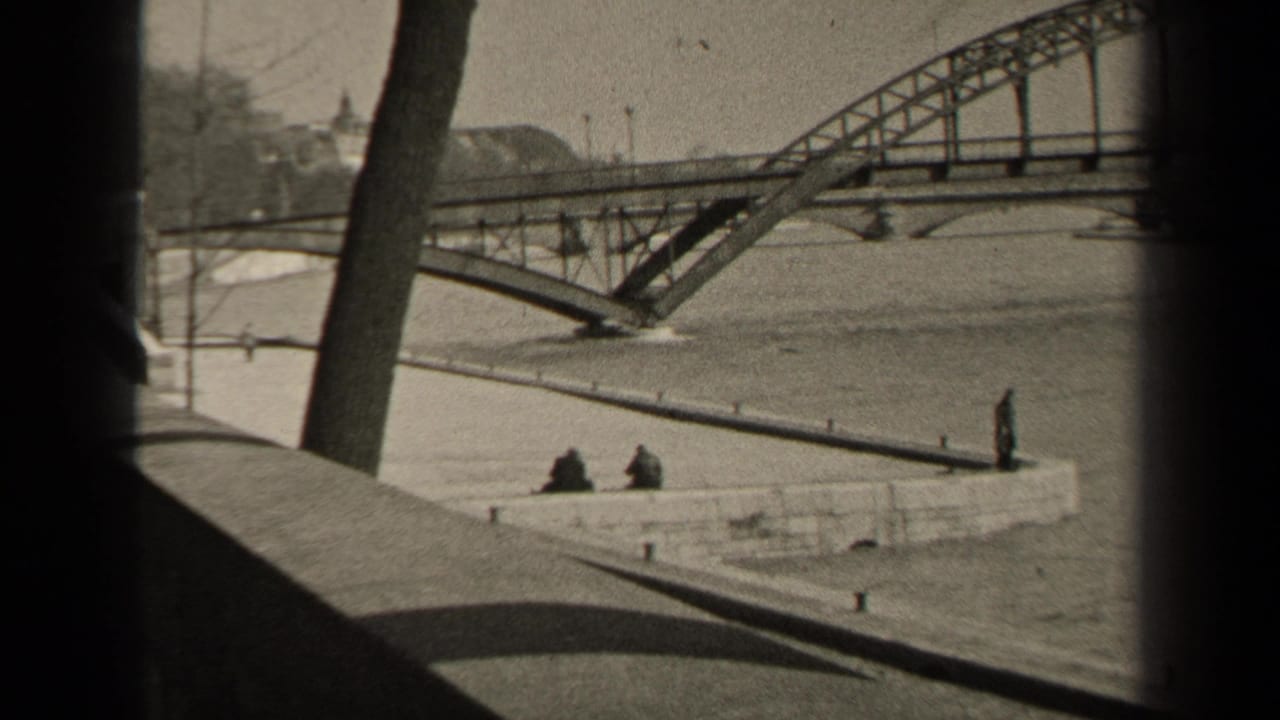 1947 Paris home-movie frame with two seated figures by the river, an arched metal bridge, bare trees, and distant buildings.