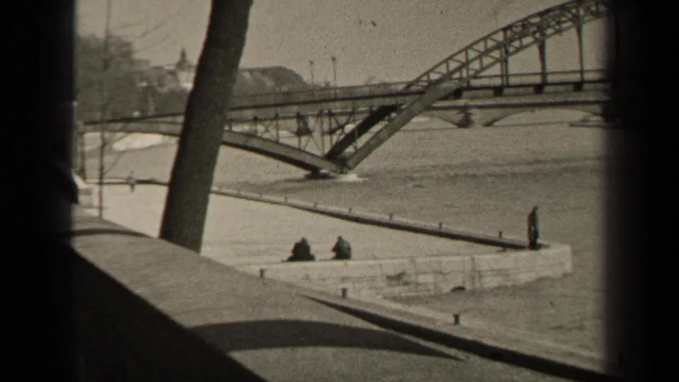 1947 Paris home-movie frame with two seated figures by the river, an arched metal bridge, bare trees, and distant buildings.
