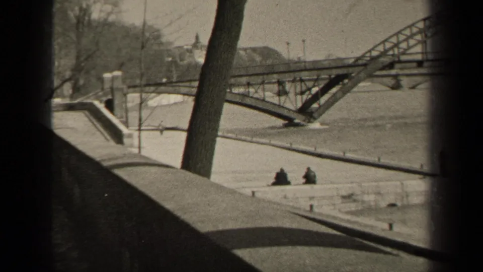 Paris riverside view with a metal bridge, stone walkway, bare trees, and two distant seated people near the water.