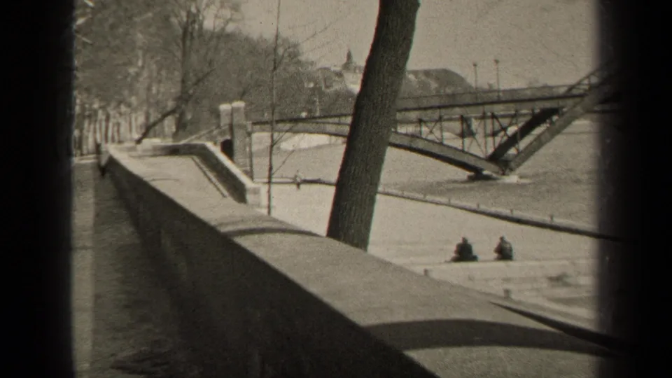1947 Paris frame showing a riverside wall, tree-lined walkway, arched bridge, water, and two seated figures in the distance.