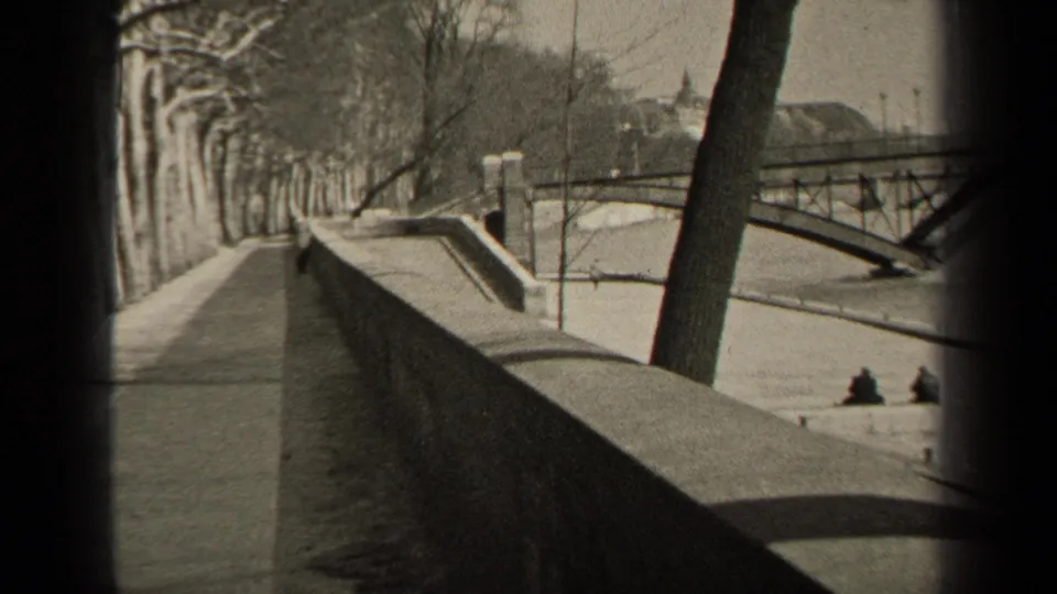Paris riverside home-movie view with a long stone wall, bare trees, arched bridge, and two distant seated people by the river.