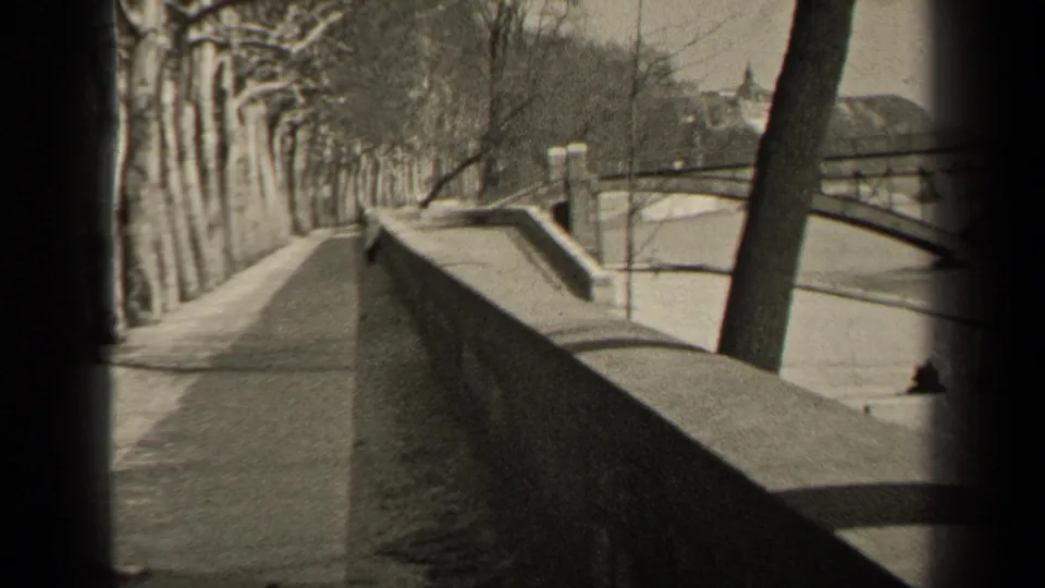 1947 Paris home-movie frame showing a bare tree-lined riverside walkway, stone embankment, water, and distant bridge-like structure.