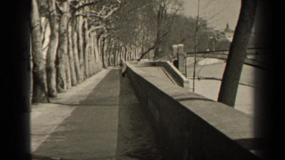 Black-and-white 8mm frame of a Paris riverside promenade with bare trees, stone wall, water, and bridge-like forms in the distance.