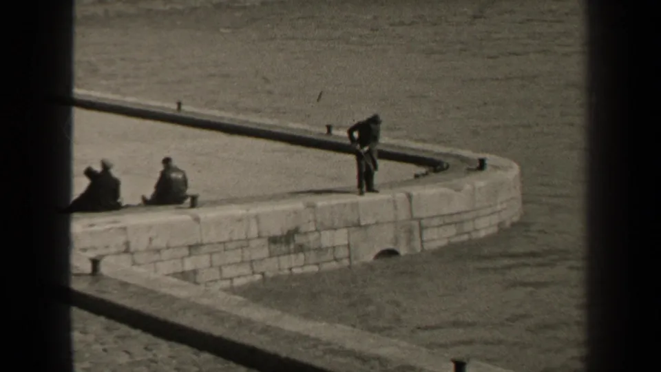 Black-and-white home-movie frame of a Paris riverside platform with seated people and one standing figure looking toward the water.