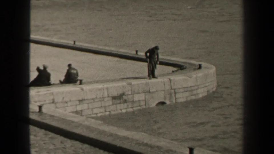 1947 Paris home-movie frame showing a man on a curved stone waterfront wall, with two seated figures nearby and water behind them.