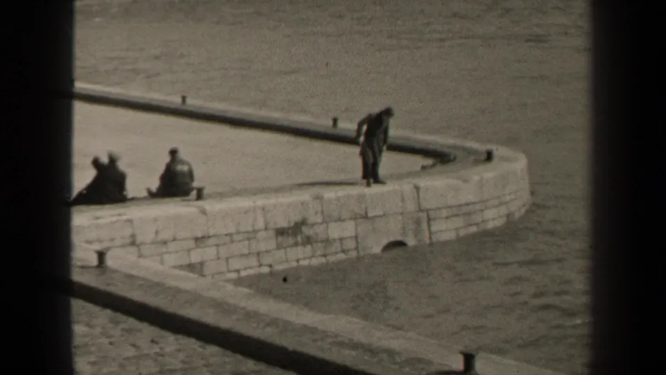 1947 Paris waterfront frame with a man bending slightly on a curved stone embankment as two people sit nearby along the river edge.
