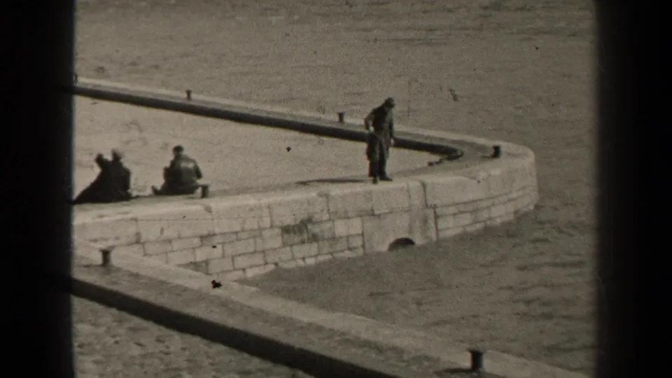 Black-and-white 8mm frame from 1947 Paris showing a man standing on a stone waterfront wall near two seated people.