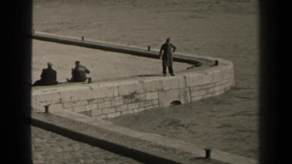 1947 Paris riverside home-movie frame with a long-coated man on the stone embankment, seated figures to the left, and water behind.