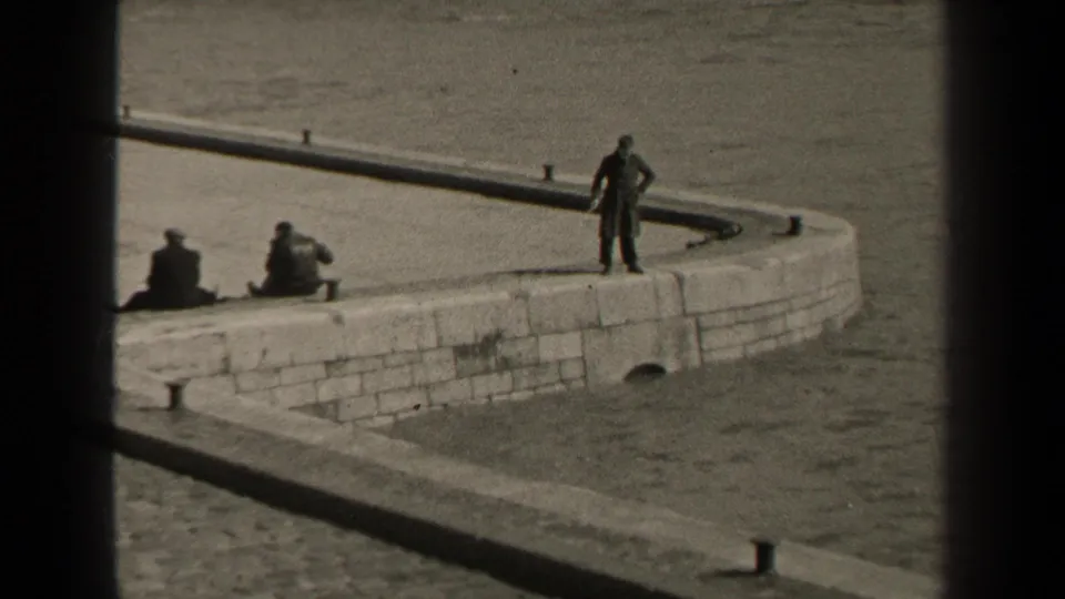 1947 Paris home-movie frame showing men on a curved stone pier by the water, including a standing fisherman and seated figures.