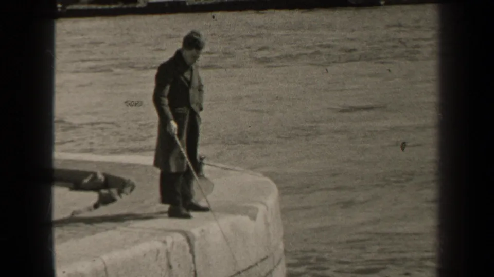 1947 Paris home-movie still of a man holding fishing gear on a stone riverside edge, with water behind him.