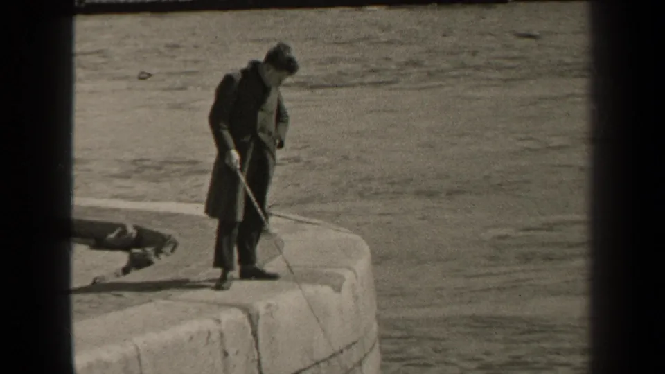 1947 Paris frame showing a man in a long coat handling his fishing line on a curved stone pier beside the water.