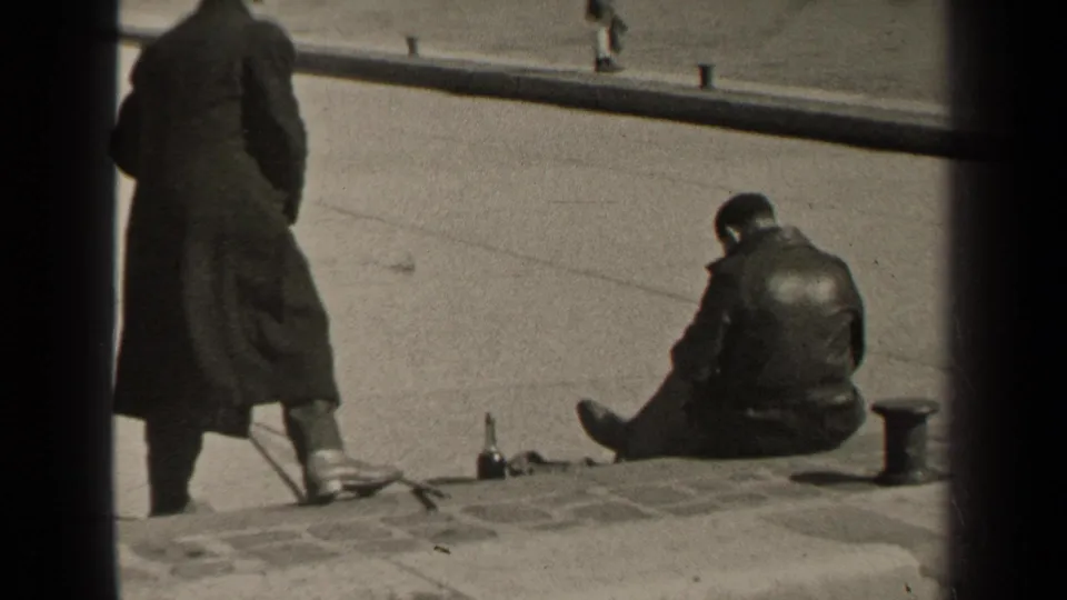 1947 Paris home-movie frame showing two men in dark outerwear on steps or curbside pavement, with one standing and one seated.