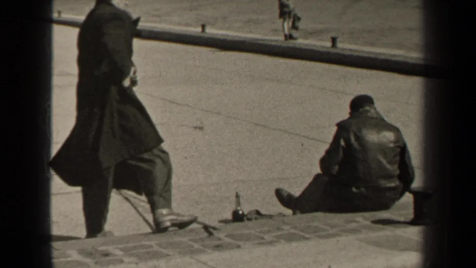 Paris 1947 home-movie still of a long-coated man standing on steps or a curb while another man sits with his back to camera.