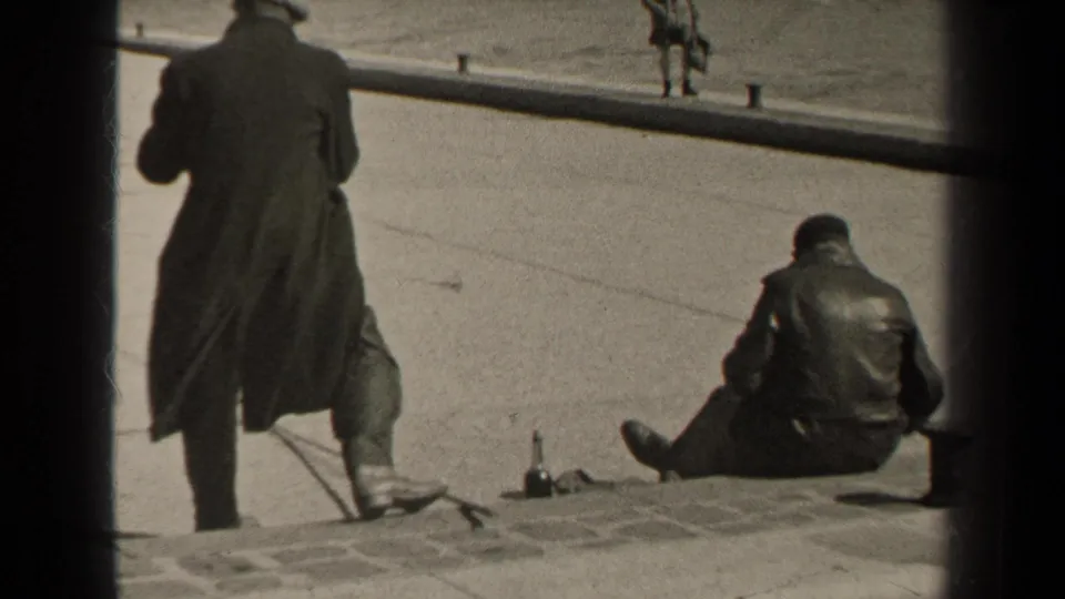 1947 Paris home-movie frame showing men in dark coats near a stone riverside embankment, one seated and one standing close by.