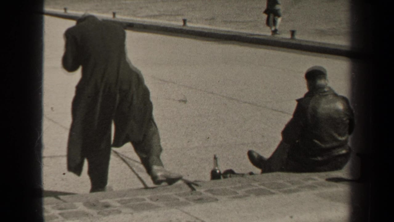 A standing man and seated man in dark coats beside a stone curb and open pavement.