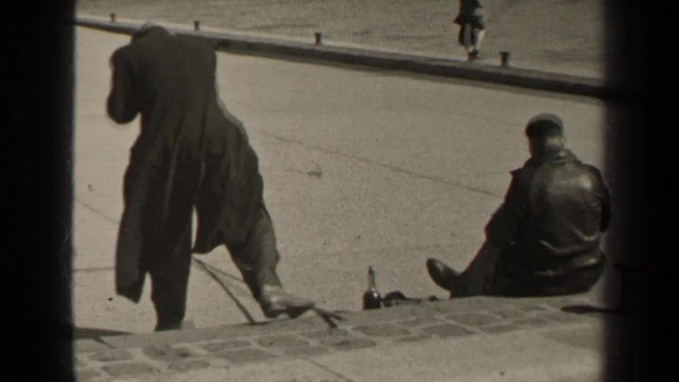 1947 Paris frame with a man in a long coat standing near a seated man on the pavement by a stone embankment.
