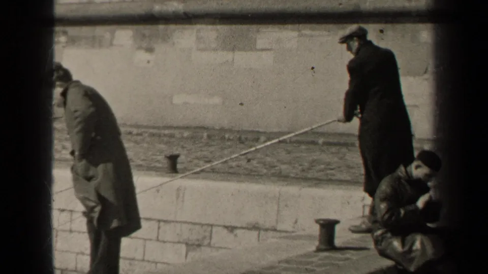 1947 Paris home-movie frame showing men near a stone embankment, including a figure holding or standing by a long pole.