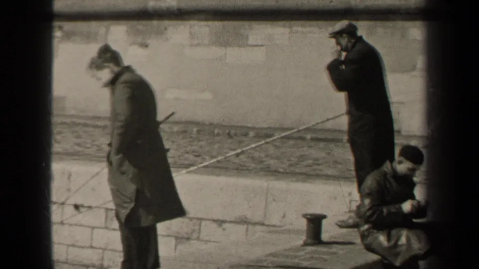 1947 Paris frame showing two standing men and a seated man beside a stone embankment, with a long pole crossing the scene.