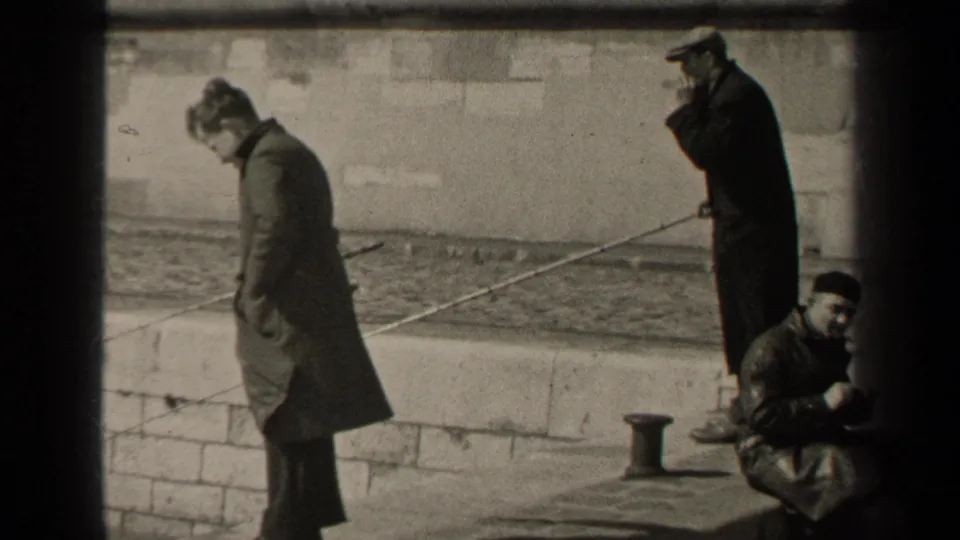 1947 Paris 8mm frame showing men in coats on a stone waterfront wall, with fishing rods, water and a seated man nearby.