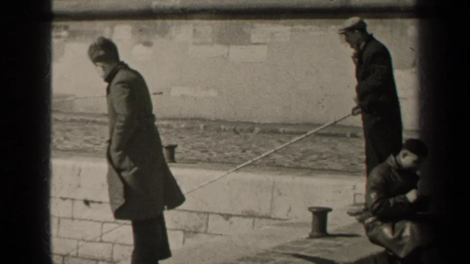 1947 Paris home-movie frame with adult men in heavy coats fishing from a stone wall beside rippled water.