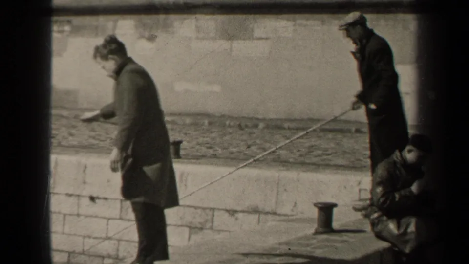 Black-and-white 8mm frame from Paris in 1947 showing men on a stone embankment, including a gesturing figure and a fisherman.