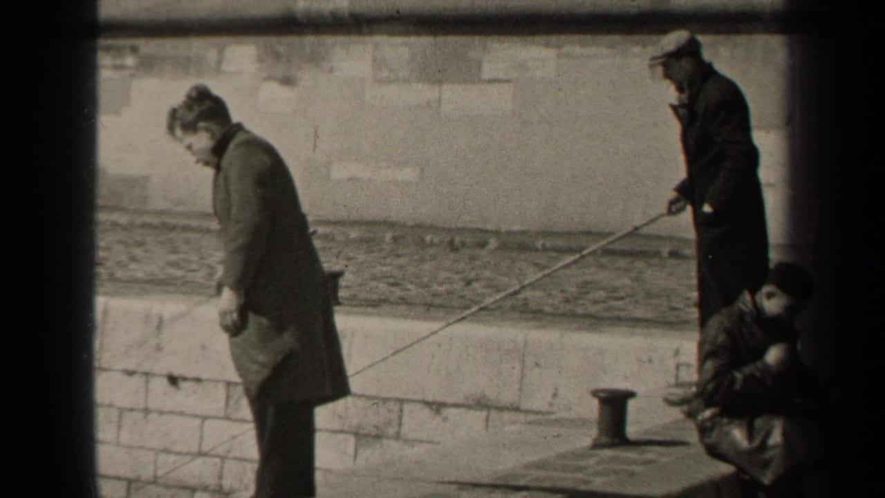 1947 Paris home-movie frame showing adult men in long coats beside a stone river wall, with fishing rods extended over the water.