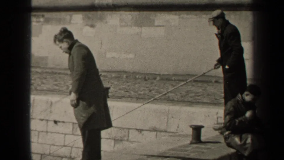 1947 Paris home-movie frame showing adult men in long coats beside a stone river wall, with fishing rods extended over the water.