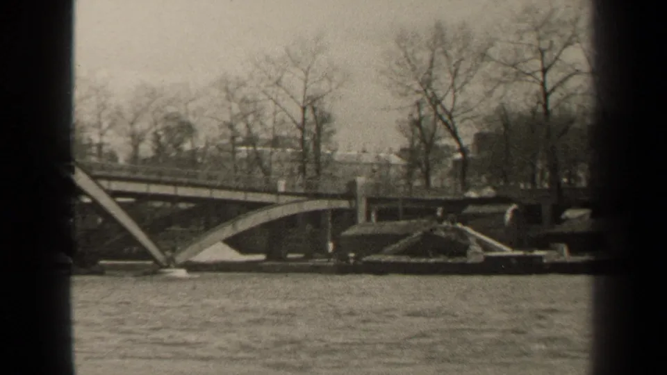 Black-and-white 1947 Paris waterfront frame showing a bridge span, bare trees, embankment details, and moving river water.