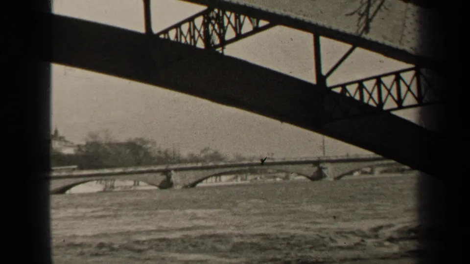 1947 Paris river view framed by dark steel trusses, with a distant stone arch bridge and bare riverbank trees.