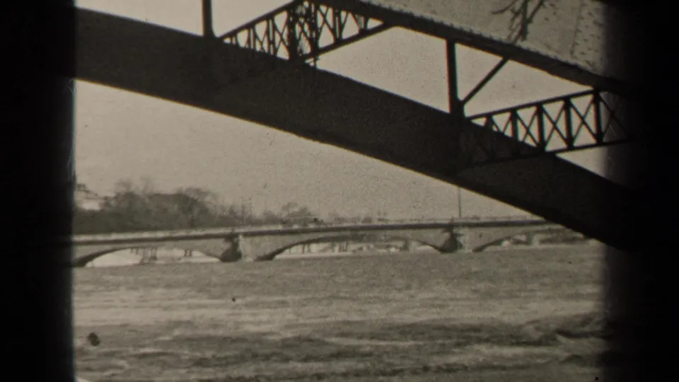 1947 Paris 8mm frame from beneath a metal bridge, with dark structural shadows over rapid water and a far stone bridge.