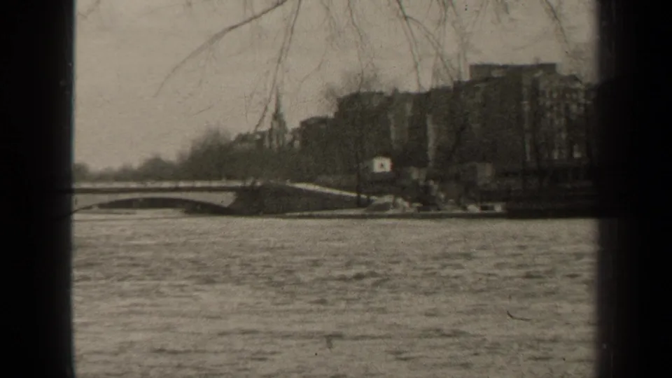 Black-and-white 8mm Paris river scene with an arched bridge, bare trees, embankment, and buildings along the far bank.