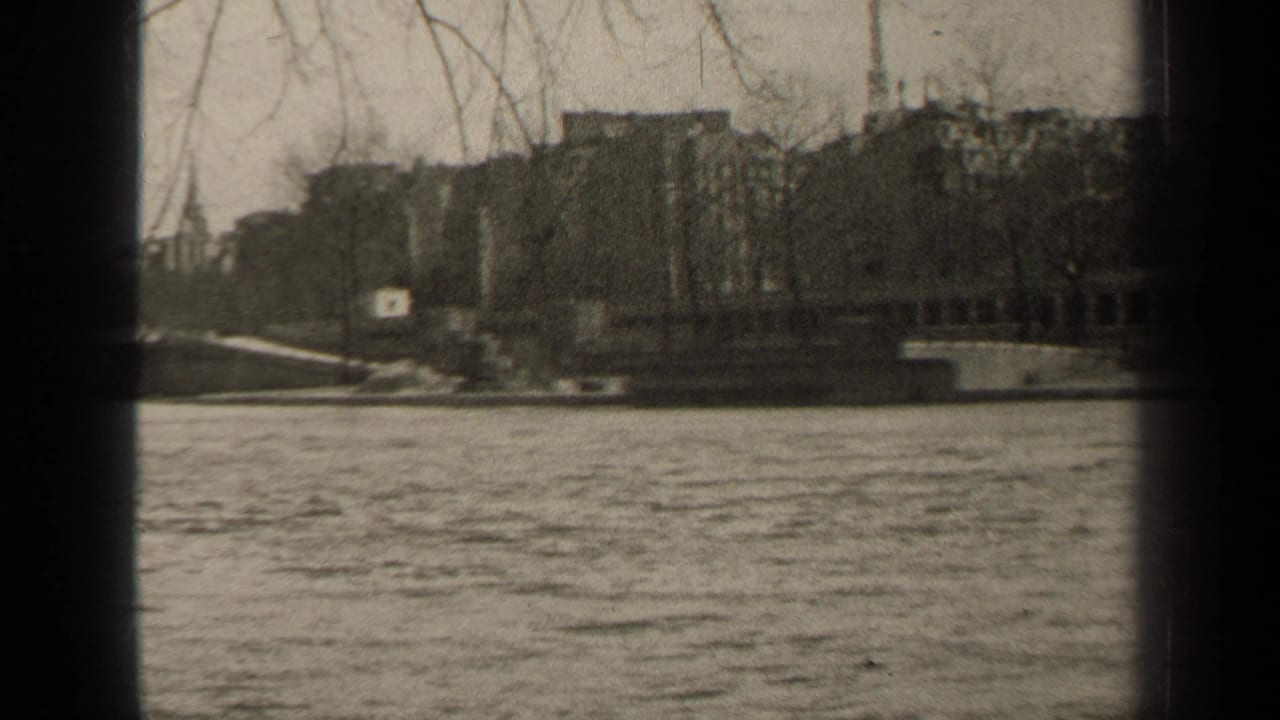 1947 Paris riverside view with water in the foreground, bare trees and buildings across the embankment, and dark 8mm frame edges.