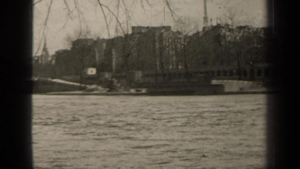 1947 Paris riverside view with water in the foreground, bare trees and buildings across the embankment, and dark 8mm frame edges.