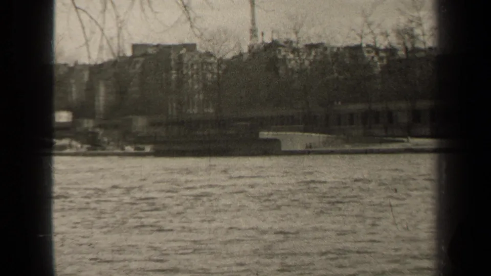 1947 Paris home-movie still looking across flowing water toward a stone embankment, bare trees, and indistinct urban buildings.