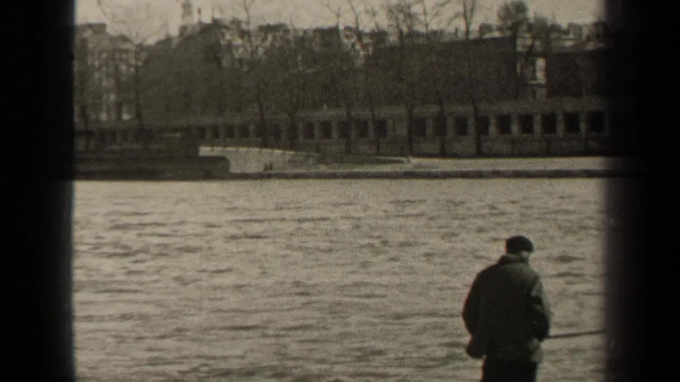 1947 Paris riverside frame with a lone man in a dark coat and hat holding a fishing rod beside the water.