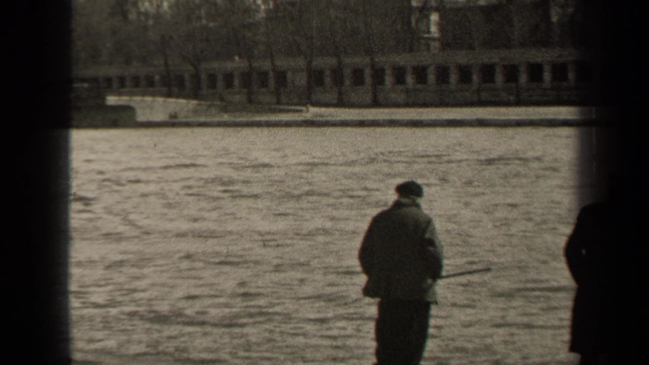 1947 Paris home-movie frame showing a man in a coat and cap holding a fishing rod near the riverbank, with water and a low bridge beyond.