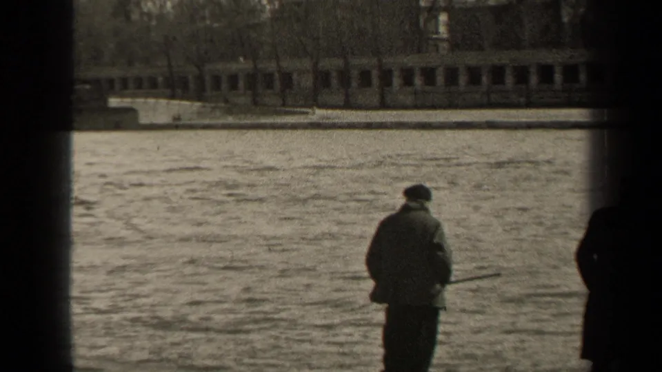 1947 Paris home-movie frame showing a man in a coat and cap holding a fishing rod near the riverbank, with water and a low bridge beyond.