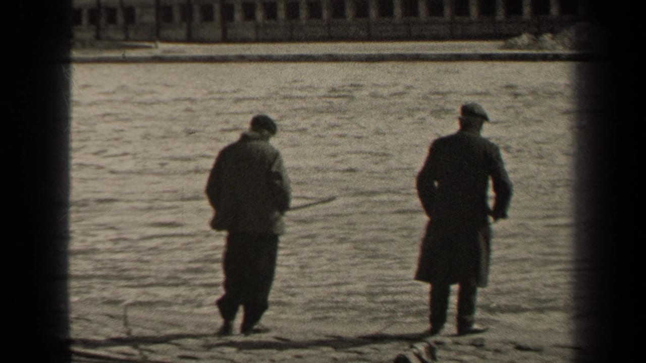 Two men in coats stand near the riverbank, one holding a fishing rod, with a bridge behind them.