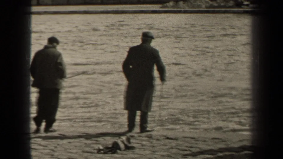 1947 Paris home-movie frame showing two men in coats moving along the riverbank, with fishing gear on the ground and water behind them.