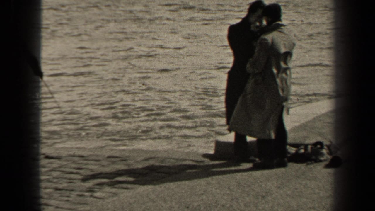 In supported 1947 Paris home-movie footage, two adult men in coats stand close on a waterside promenade with gear at their feet.