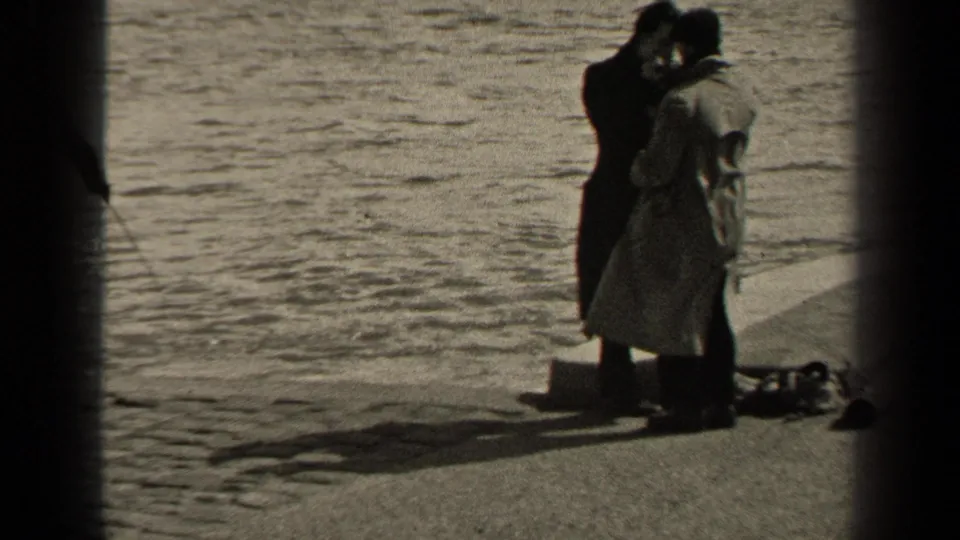 In supported 1947 Paris home-movie footage, two adult men in coats stand close on a waterside promenade with gear at their feet.