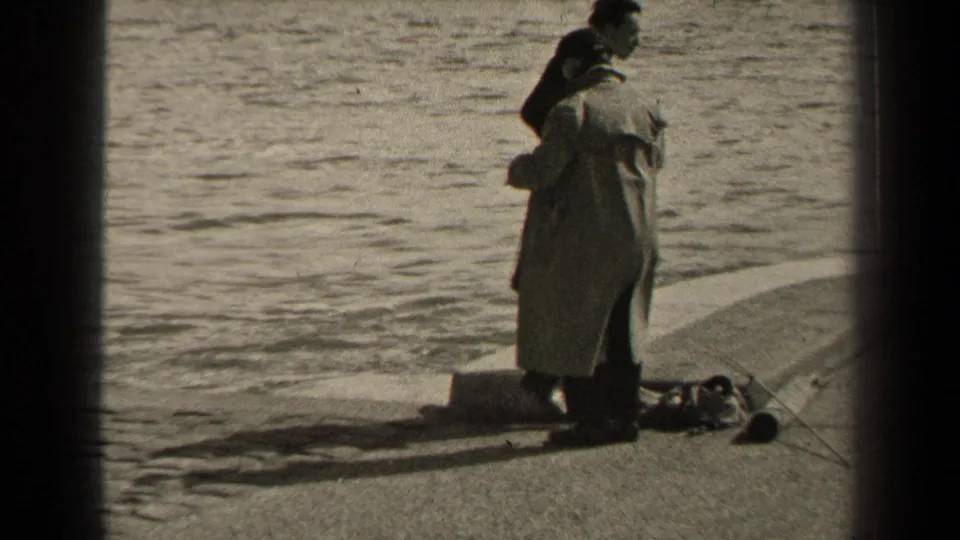 A man in a light coat faces the water while another adult stands close behind him on the Paris promenade, with tackle at their feet.