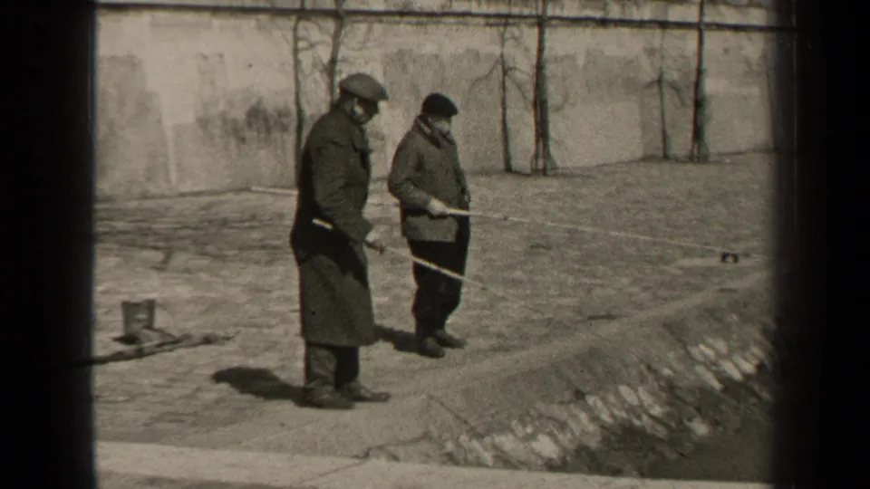 Two adult men in coats handle fishing rods on a Paris waterside embankment, with a low wall and bare vines behind them.
