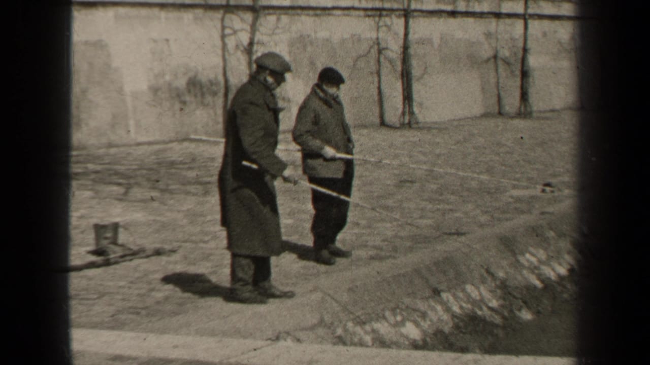 In 1947 Paris home-movie footage, two adult men in winter coats and hats stand on paved ground beside a wall, holding long poles.
