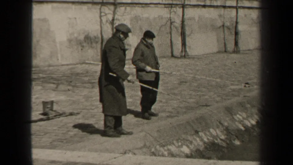 In 1947 Paris home-movie footage, two adult men in winter coats and hats stand on paved ground beside a wall, holding long poles.