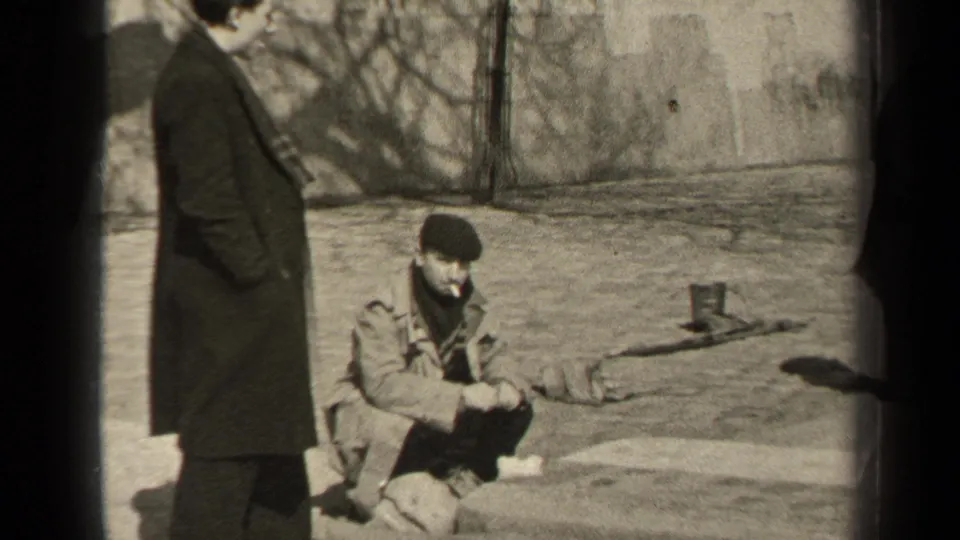 In the Paris 1947 home-movie frame, one man stands partly cropped at left as another sits on the pavement in a beret and smokes.