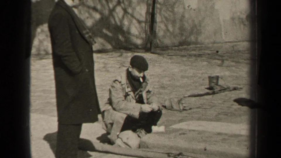 In 1947 Paris, two adult men pause outdoors in winter coats, with one crouched on a stone ledge smoking while another stands nearby.