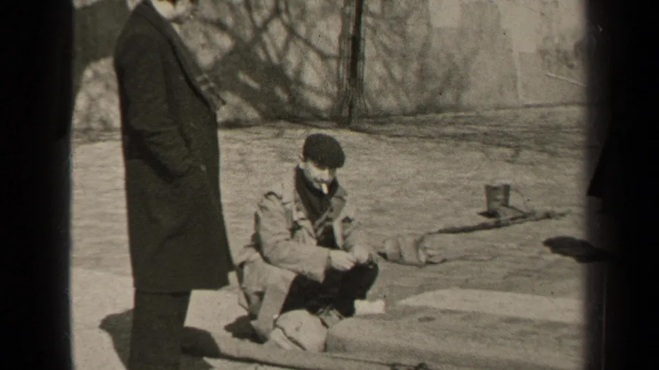 In the Paris home-movie scene, a man stands at left while another crouches on a ledge with a cigarette, framed by bare branches and pavement.