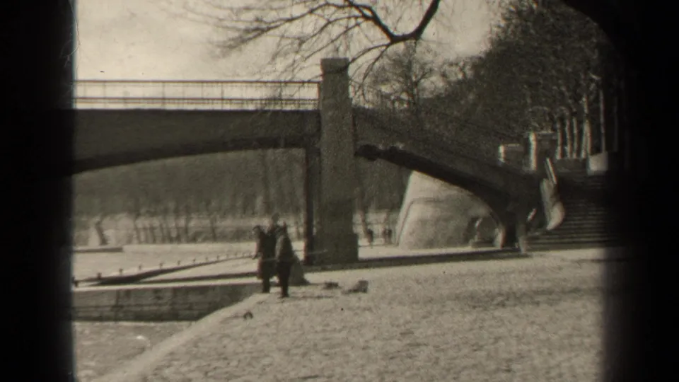 A Paris riverside view shows a bridge over water, steps, winter trees, and small figures walking along the embankment.